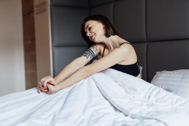 Mulher morena de cabelos longos felizes na cama branca na luz suave da manhã sob o edredom