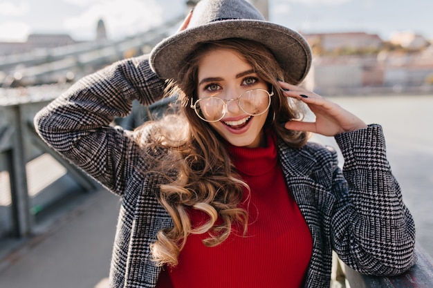 Mulher morena de olhos azuis com expressão facial feliz posando com prazer em desfocar o fundo da cidade em dia de outono