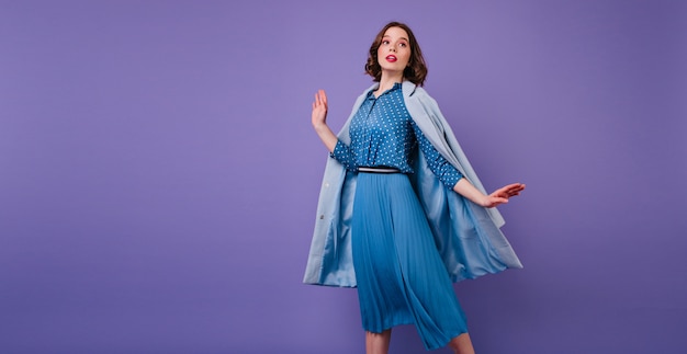 Mulher morena emocional com casaco azul, posando na parede roxa. Foto interna da bela modelo feminina de cabelos curtos em um vestido midi na moda.