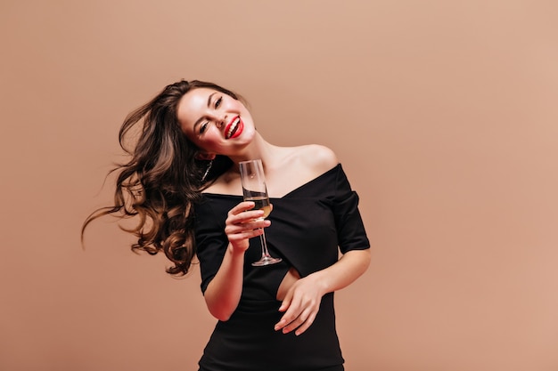 Mulher morena encantadora com lábios vermelhos é linda, sorrindo e brincando com o cabelo. menina com roupa preta, posando com taça de champanhe.