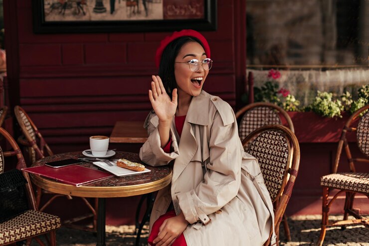 Foto grátis mulher morena feliz com vestido vermelho de trench coat e boina estilosa acena com a mão em saudação sorri amplamente e senta-se no café da rua