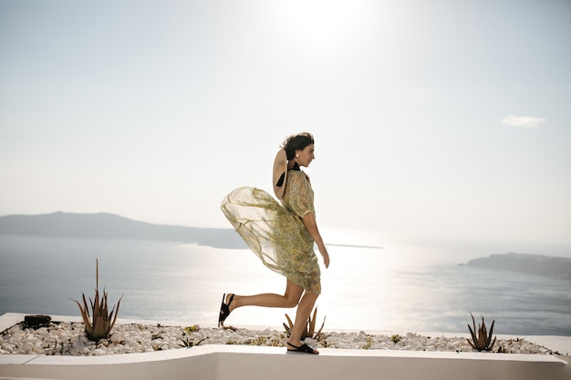Foto grátis mulher morena feliz em velejador e vestido floral se move no fundo do mar encantadora senhora encaracolada com roupa elegante caminha no terraço com vista para o mar