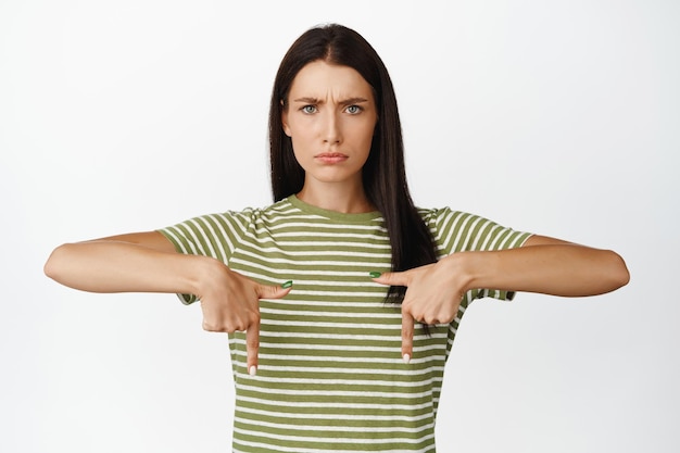 Mulher morena irritada franzindo a testa e emburrada apontando dedos para cima em smth mau pé na camiseta sobre fundo branco