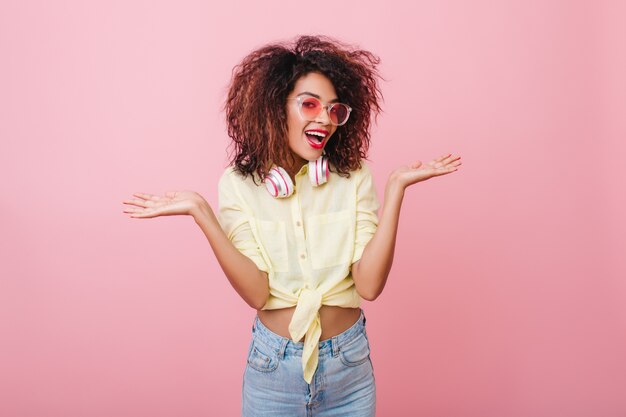 Mulher morena maravilhosa com penteado encaracolado, posando com um sorriso gentil na camisa amarela da moda. Retrato interior de garota africana elegante confiante com cabelo castanho escuro.