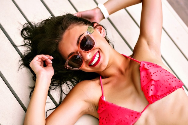 Mulher morena muito alegre positiva sorrindo e rindo, tendo um grande momento sozinho, deitado no chão de madeira perto da piscina, semana para biquíni e óculos de sol, férias, férias, tempo.