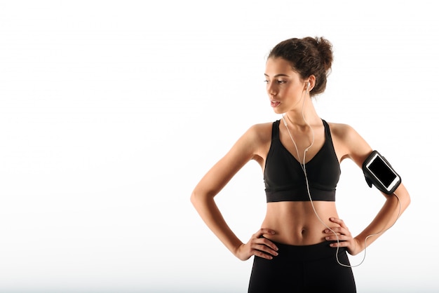 Mulher morena muito encaracolado fitness segurando os braços nos quadris e ouvir música enquanto olhando para longe