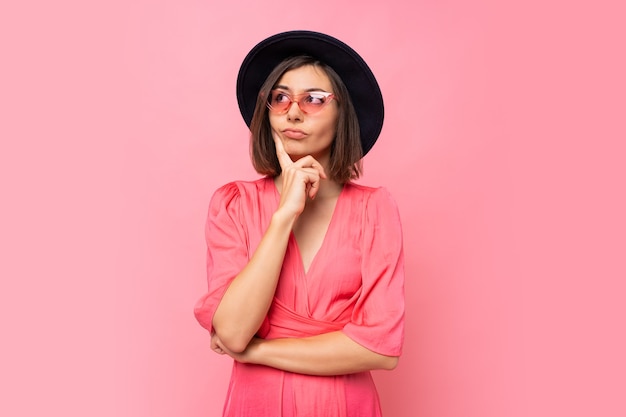 Mulher morena pensativa em óculos elegantes, posando sobre parede rosa.