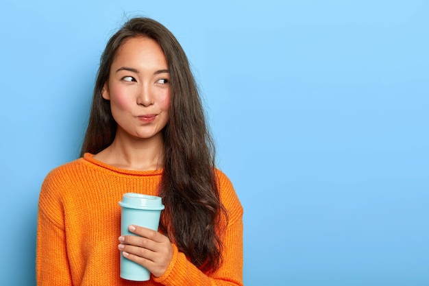 Mulher morena pensativa franze os lábios, olha pensativamente para o lado, segura o café para viagem, toma uma decisão em mente, planeja o dia, usa suéter laranja, fica de pé sobre a parede azul