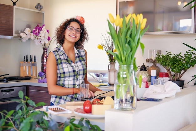 Mulher morena positiva com cabelo encaracolado faz salada com tomate e batata em uma cozinha em casa.