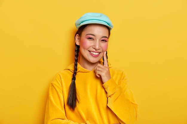 Mulher morena positiva com pele saudável, tocando a bochecha ruge com o dedo indicador, sorrindo feliz, vestida com um moletom amarelo casual