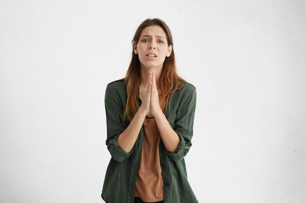 Foto grátis mulher morena preocupada e apologética tendo algumas dificuldades para pressionar as palmas das mãos, implorando por desculpas. mulher apreensiva expressando sua ansiedade pedindo a deus por saúde isolada sobre parede branca