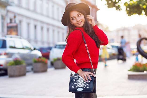Foto grátis mulher morena sedutora em roupa casual outono, andando na cidade ensolarada. pulôver de malha vermelho, chapéu preto moderno, saia de couro.