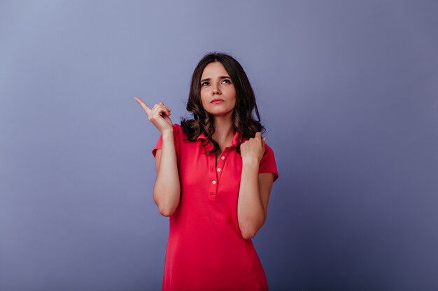 Mulher morena séria em vestido vermelho casual, olhando para cima. retrato de menina glamorosa pensativa isolada na parede roxa.