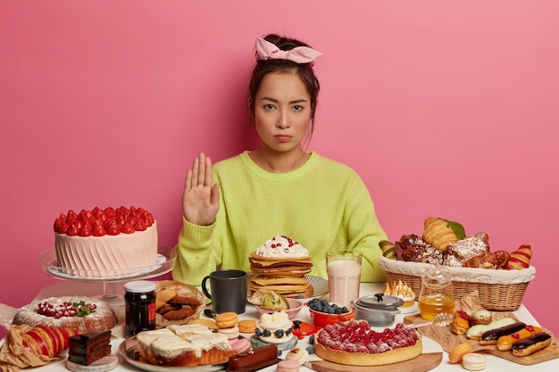 Foto grátis mulher morena séria faz gesto de recusa, posa perto da mesa da cozinha com sobremesas, mantém a dieta, recusa comer alimentos com muitas calorias. doces da moda