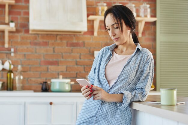 Mulher morena séria manda mensagens online, usa conexão gratuita à Internet, usa camisa casual,