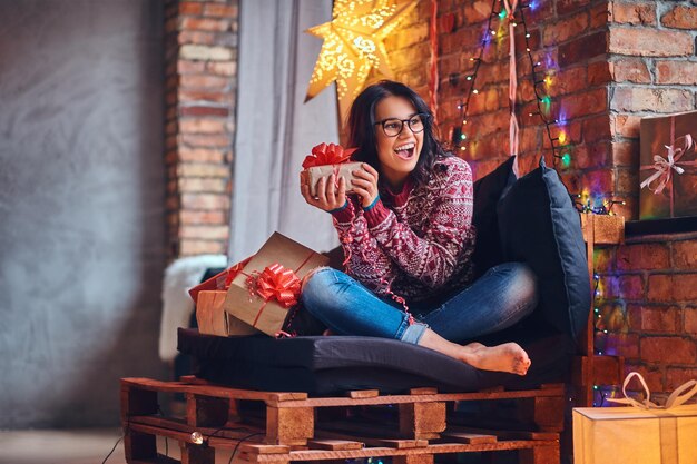 Mulher morena sexy com pé nu, vestida de jeans e um suéter vermelho, posando em um sofá em uma sala com decoração de Natal.