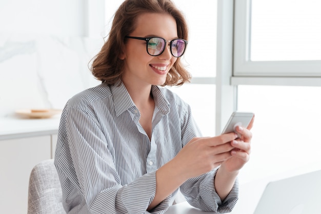 mulher morena sorridente em copos mandando mensagem no smartphone enquanto está sentado na cozinha