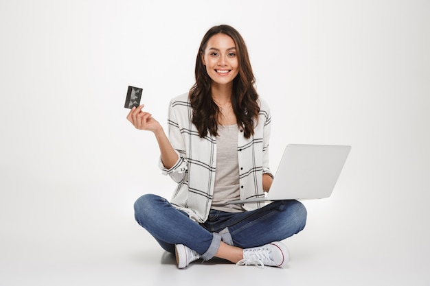 Mulher morena sorridente na camisa, sentada no chão com o computador portátil e cartão de crédito enquanto olha para a câmera sobre cinza