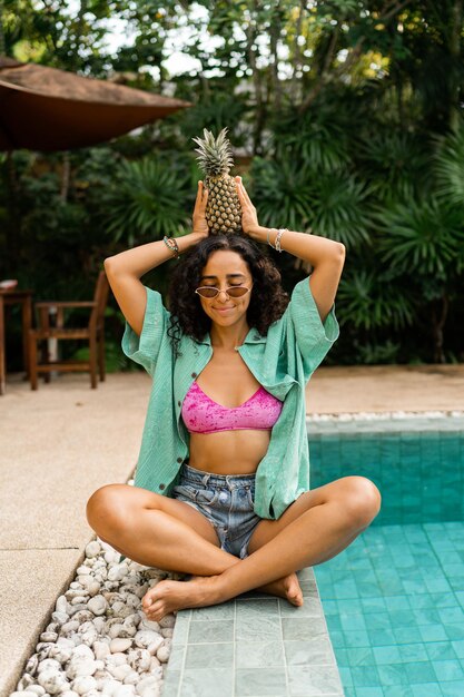 Mulher morena sorridente posando com abacaxi sentado perto da piscina em resort tropical