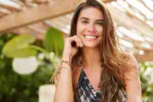 Foto grátis mulher morena sorridente sentada em um refeitório ao ar livre, tem um sorriso largo, tem bom descanso, tem expressão satisfeita, descansa em um resort tropical. jovem bonita descansa no terraço do café. positividade