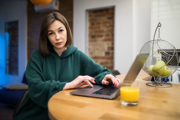 Mulher morena surpresa está trabalhando em seu laptop na mesa da cozinha, bebendo suco de laranja
