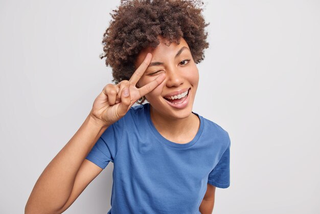 mulher mostra o sinal da paz sobre os olhos piscadelas e sorrisos usa camiseta azul casual isolada no branco demonstra gesto de vitória aproveita a vida. Conceito de linguagem corporal