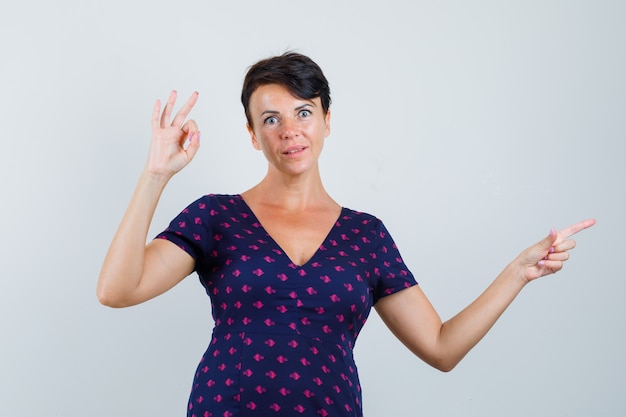 Mulher mostrando gesto de ok, apontando para o lado no vestido e parecendo espantada