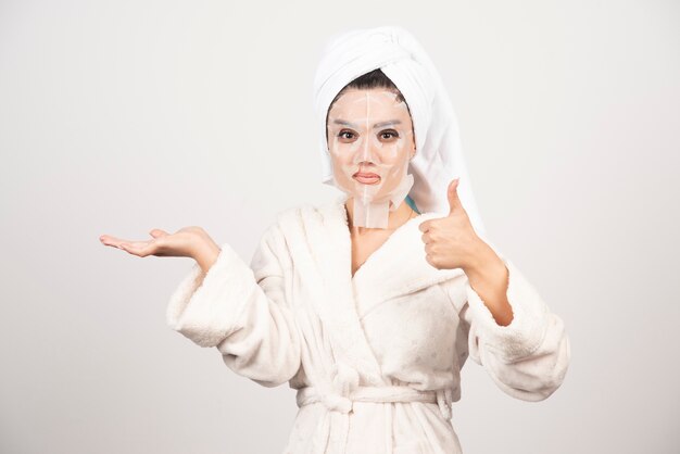 Mulher mostrando o polegar para cima, vestindo roupão e toalha com máscara