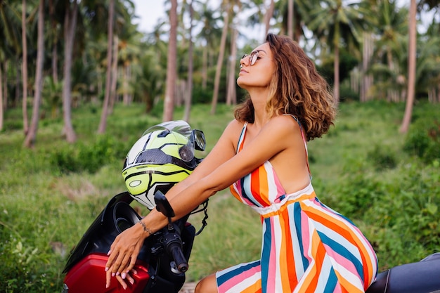 Mulher motociclista caucasiana feliz com vestido colorido de verão de férias com capacete de motociclista