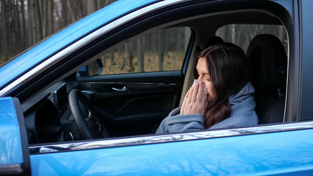 Mulher motorista com resfriado e com nariz escorrendo tosse e assoa o nariz em guardanapo de papel sentado no volante do carro na estrada