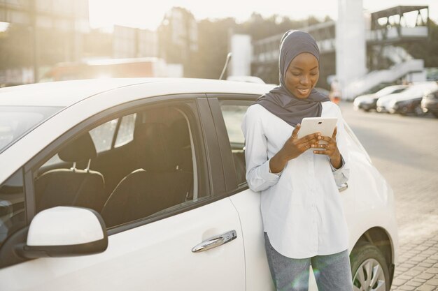 Mulher muçulmana africana encostada no carro dela e segurando um tablet digital. Trabalhando remotamente ou compartilhando informações.