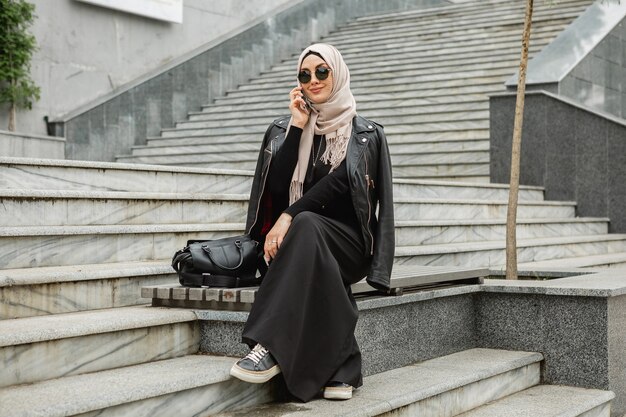 Mulher muçulmana elegante e moderna em hijab, jaqueta de couro e abaya preta andando na rua da cidade falando em smartphone