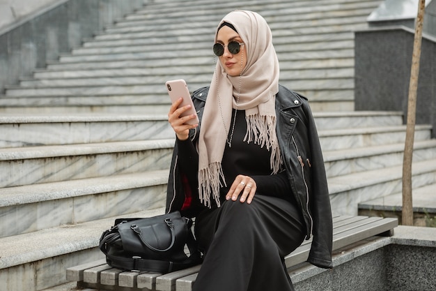 Mulher muçulmana elegante e moderna em hijab, jaqueta de couro e abaya preta andando na rua da cidade usando smartphone