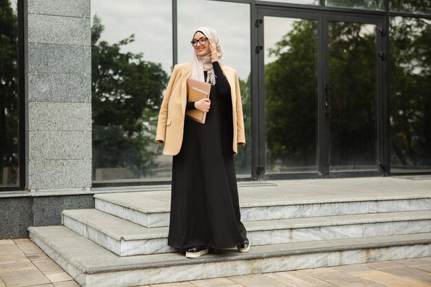 Mulher muçulmana elegante e moderna em hijab, jaqueta estilo empresarial e abaya preta andando na rua da cidade com laptop