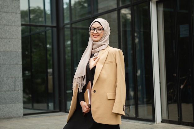 Mulher muçulmana elegante e moderna em hijab, jaqueta estilo empresarial e abaya preta andando na rua da cidade com laptop