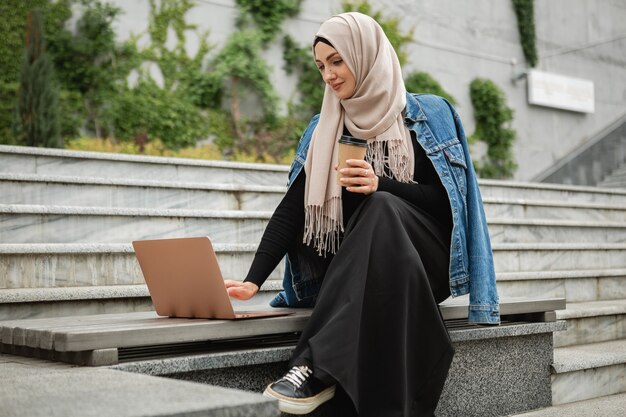 Mulher muçulmana elegante e moderna em hijab, jaqueta jeans e abaya preta sentada na rua da cidade trabalhando em um laptop