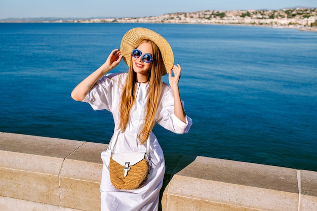 Foto grátis mulher muito elegante com vestido branco, chapéu de palha e bolsa posando perto do mar