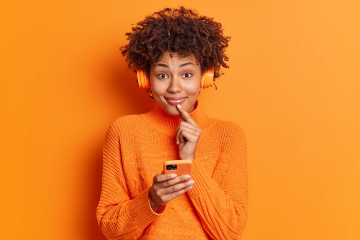 Foto grátis mulher muito satisfeita sorri suavemente na frente mantém o dedo perto dos lábios usa smartphone para enviar mensagens de texto online e ouvir música usa fones de ouvido sem fio estéreo