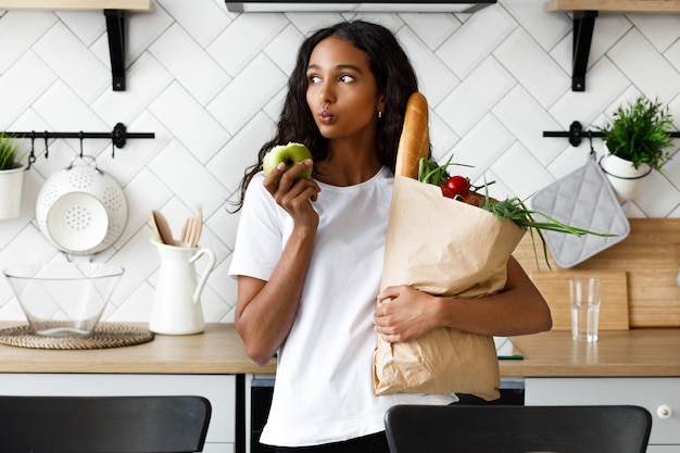 Mulher mulata pensativa está segurando o pacote cheio de legumes frescos em uma mão e maçã mordida na outra, na moderna cozinha branca