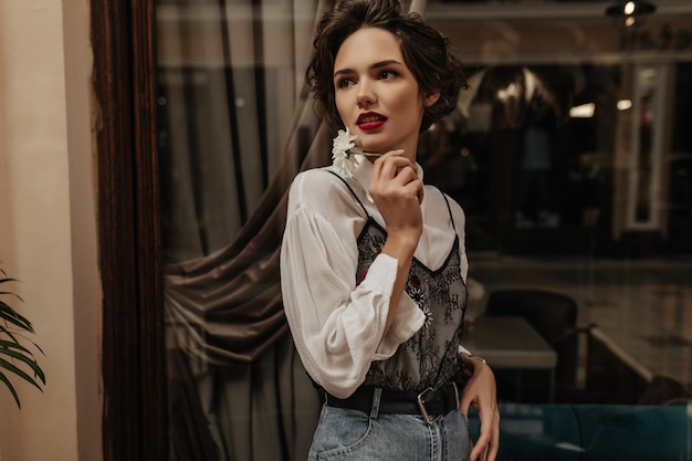 Mulher na moda em jeans com cinto e camisa branca, segurando uma flor dentro. Mulher moderna com penteado curto e poses de lábios brilhantes no café.