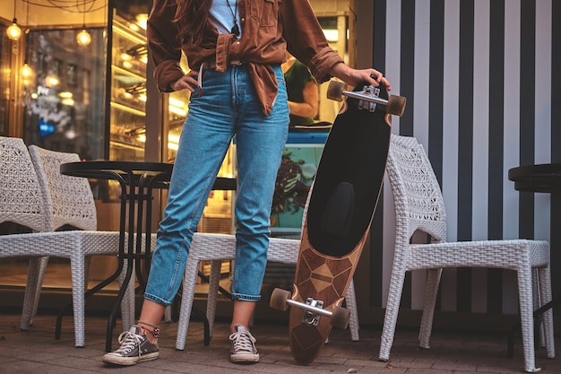 Mulher na moda hipster está posando para o fotógrafo com seu longboard perto do café.