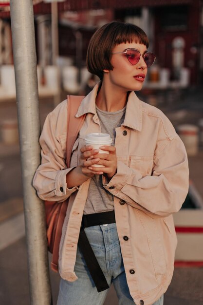 Mulher na moda na jaqueta leve segura xícara de café ao ar livre Garota de cabelos curtos em óculos de sol brilhantes e jeans com mochila marrom posando na cidade