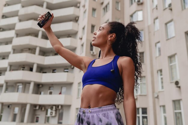 Mulher negra afro-americana atraente em roupa urbana de fitness esportiva no telhado fazendo malhar estilo de cabelo encaracolado confiante corpo atlético usando smartphone tirando foto de selfie