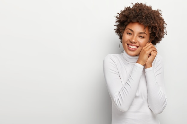 Mulher negra alegre com sorriso agradável, mantém as mãos juntas perto do rosto, parece feliz