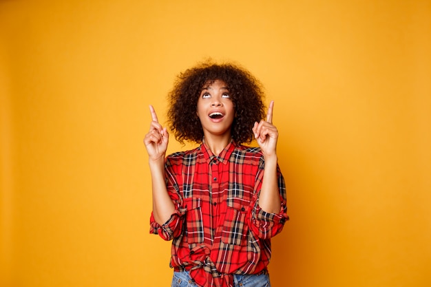 Mulher negra americana alegre na camisa vermelha que olha para cima e que aponta os dedos acima no espaço da cópia isolado sobre o fundo alaranjado.