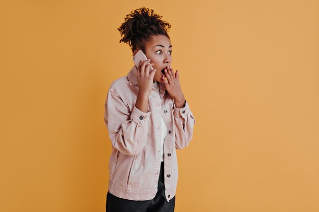 Mulher negra chocada falando no smartphone Foto de estúdio de uma garota afro-americana surpresa isolada em fundo laranja