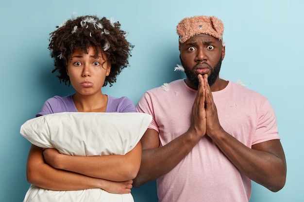 Mulher negra deprimida com corte de cabelo afro, abraça travesseiro branco, negro chocado mantém as palmas das mãos juntas, tem olhos esbugalhados, usa máscara de dormir, fica perto um do outro. Conceito de sono e descanso