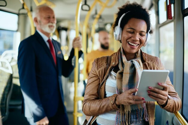 Mulher negra feliz com fones de ouvido com chamada de vídeo pelo touchpad enquanto se desloca de ônibus