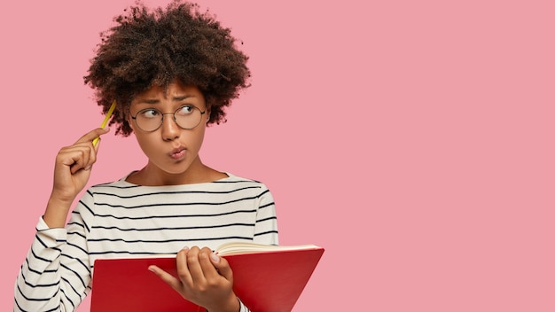 Foto grátis mulher negra perplexa tem expressão contemplativa, escreve para fazer lista de metas, segura caderno, coça a cabeça com lápis