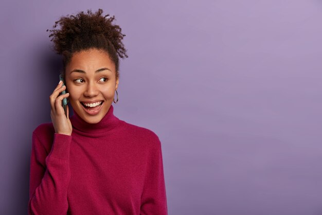 Mulher negra satisfeita conversa ao telefone, olha para o lado, discute notícias agradáveis com um amigo próximo, liga para alguém, usa gola rulê bordô